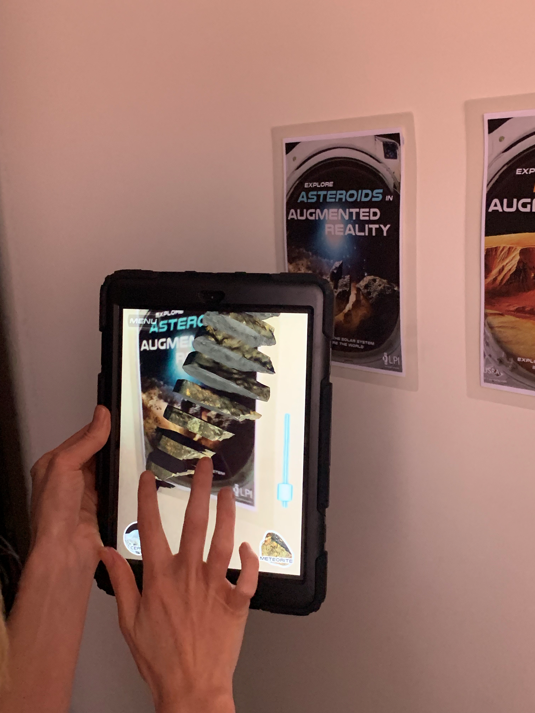 Photograph of a person holding up an iPad to a poster on the wall. The iPad shows a 3D version of the image on the wall.
